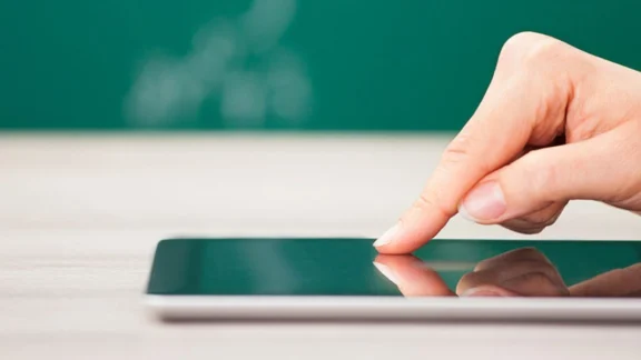 Vor einer Tafel liegt ein Handy auf einem Tisch. Eine Hand wischt über den Bildschirm. Link zum Projekt "Themendossiers für den digitalen Unterricht".