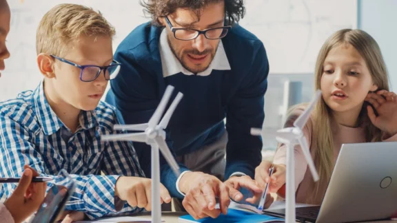 Ein Lehrer ist über einen Tisch gebeugt, an dem mehrere Kinder sitzen. Auf dem Tisch stehen kleine Windrad-Modelle. Link zum Projekt "Energie macht Schule".