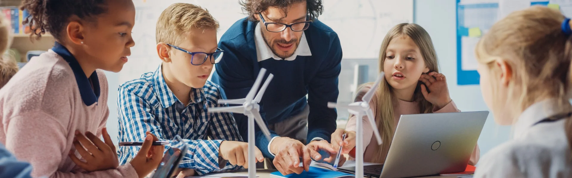 Ein Lehrer ist über einen Tisch gebeugt, an dem mehrere Kinder sitzen. Auf dem Tisch stehen kleine Windrad-Modelle. Link zum Projekt