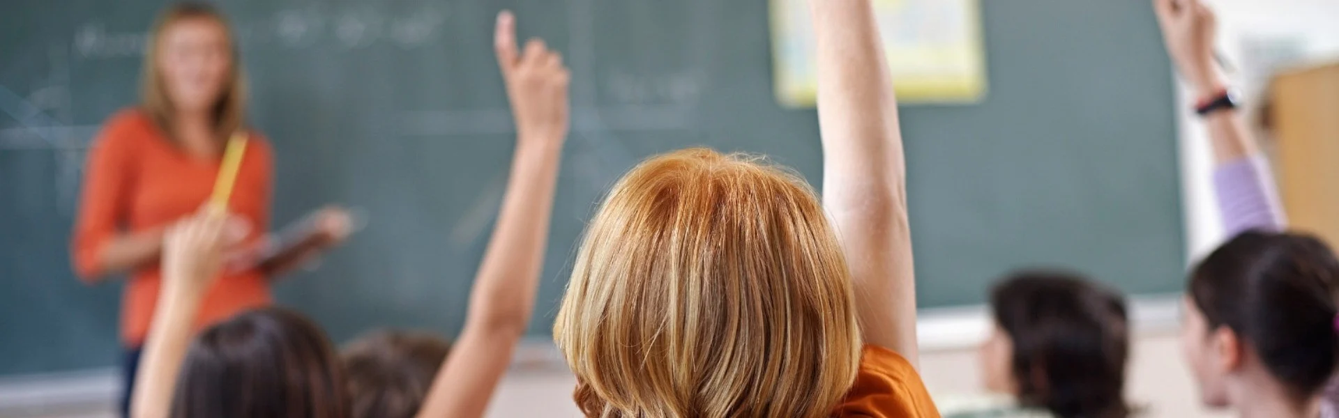 Kinder sitzen in einem Klassenraum und melden sich. An der rechten Seite steht